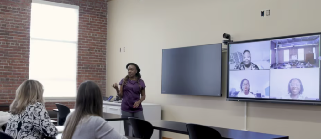 Instructor standing at the front of a classroom facing a group of students in front of her and another group of students virtually on a large TV screen behind her.