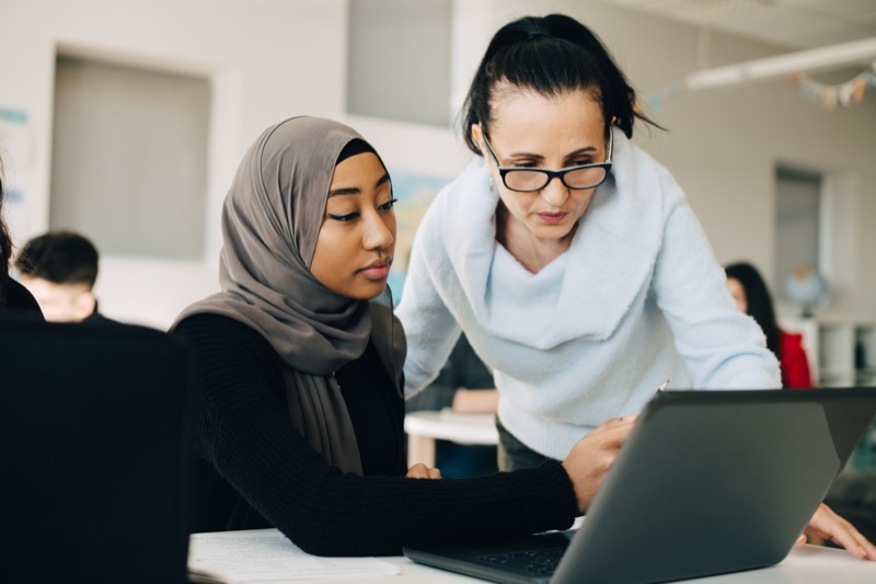 An instructor and student collaborate on a laptop with guidance from a supportive teacher, embodying the essence of great teaching in a diverse classroom setting.