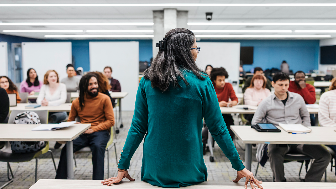 Teacher stands at the front of her classroom and faces here students.
